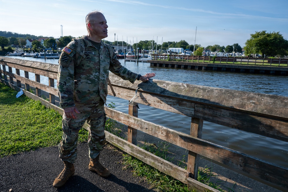 A U.S. Army soldier in MultiCam/OCP camouflage uniform stands along a waterfront area next to a wooden fence, appearing to gesture or point toward the water. The background shows a marina with sailboats and motorboats docked along a pier, surrounded by trees and green vegetation. The setting suggests a post-event assessment or inspection of a waterfront location.