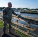 A U.S. Army soldier in MultiCam/OCP camouflage uniform stands along a waterfront area next to a wooden fence, appearing to gesture or point toward the water. The background shows a marina with sailboats and motorboats docked along a pier, surrounded by trees and green vegetation. The setting suggests a post-event assessment or inspection of a waterfront location.