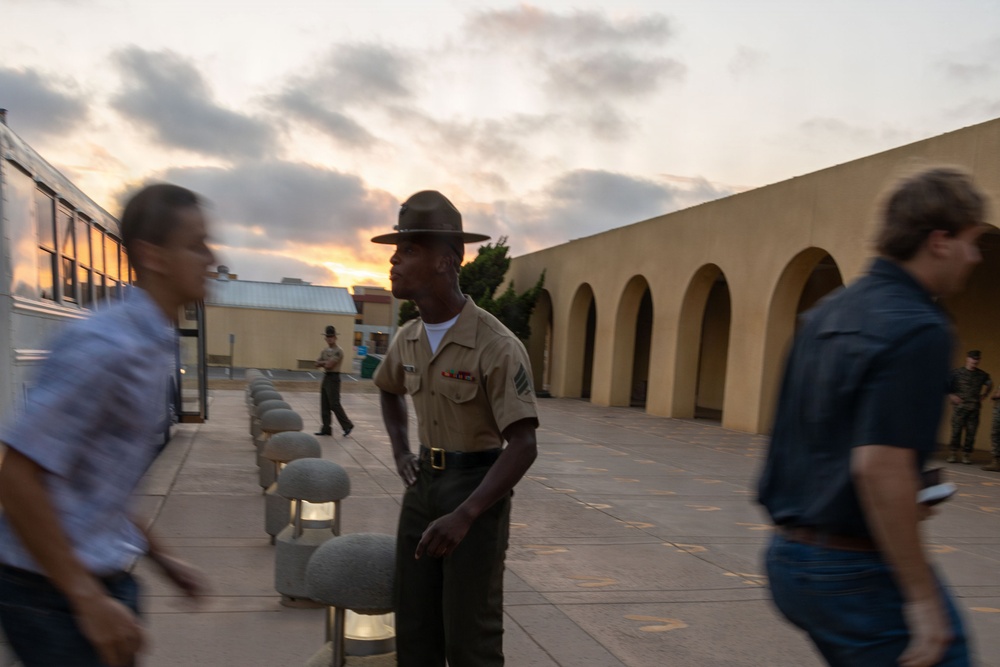 A U.S. Marine in dress uniform, wearing a drill instructor's campaign cover (campaign hat) and displaying rank insignia, stands at attention in an outdoor courtyard area at dusk. Two civilians in motion blur pass on either side of the Marine, suggesting arrival or movement through the area. The setting appears to be a military installation, likely MCRD (Marine Corps Recruit Depot) based on the distinctive arched architecture.