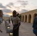 A U.S. Marine in dress uniform, wearing a drill instructor's campaign cover (campaign hat) and displaying rank insignia, stands at attention in an outdoor courtyard area at dusk. Two civilians in motion blur pass on either side of the Marine, suggesting arrival or movement through the area. The setting appears to be a military installation, likely MCRD (Marine Corps Recruit Depot) based on the distinctive arched architecture.
