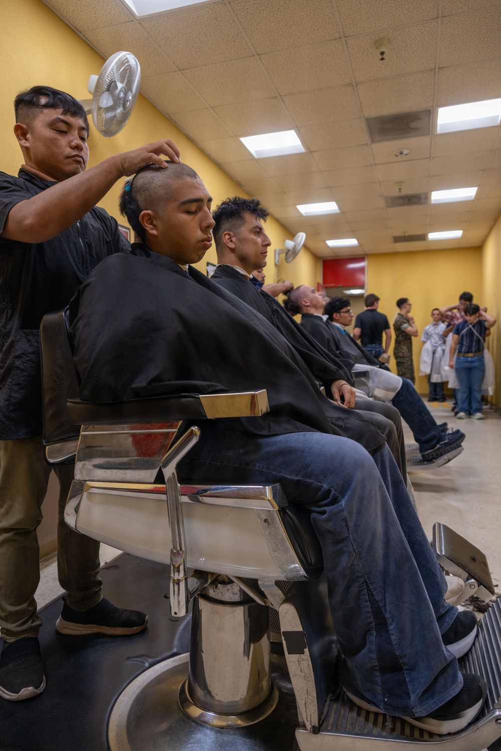 A busy barbershop scene showing a row of young men seated in classic chrome barber chairs receiving haircuts simultaneously. The foreground subject is receiving a close-cropped or shaved head cut, consistent with military induction processing. The long room with yellow walls contains multiple barbers and clients extending into the background.
