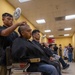 A busy barbershop scene showing a row of young men seated in classic chrome barber chairs receiving haircuts simultaneously. The foreground subject is receiving a close-cropped or shaved head cut, consistent with military induction processing. The long room with yellow walls contains multiple barbers and clients extending into the background.
