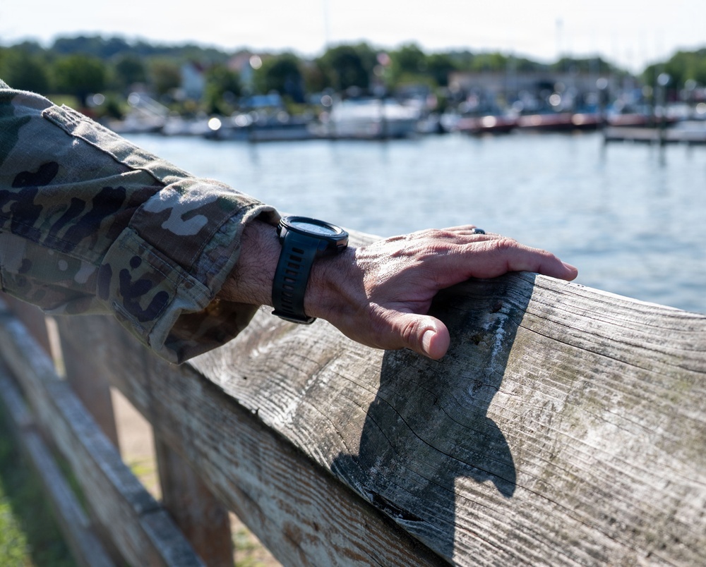 A person wearing a military camouflage uniform rests their hand on a weathered wooden fence or railing near a waterfront. A black tactical/sports watch is visible on their wrist, and a ring is visible on one finger. The background shows a marina or harbor with boats, trees, and buildings in soft bokeh focus.