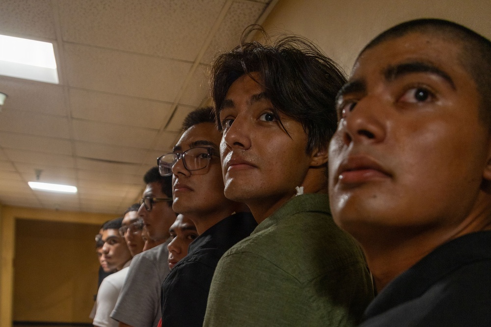 A group of young men are standing in a line inside an indoor facility with fluorescent lighting. They appear to be looking in the same direction, possibly at something or someone off-camera. The image is taken from a low angle, emphasizing the faces of the individuals in the foreground.