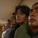 A group of young men are standing in a line inside an indoor facility with fluorescent lighting. They appear to be looking in the same direction, possibly at something or someone off-camera. The image is taken from a low angle, emphasizing the faces of the individuals in the foreground.