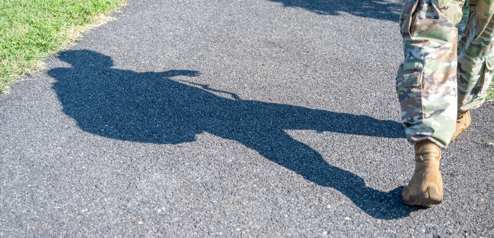 The image shows the lower body of a person wearing military camouflage uniform and tan combat boots walking on an asphalt path. A prominent shadow is cast on the pavement, revealing the silhouette of the full figure including what appears to be a helmet or headgear and possibly carried equipment. Green grass is visible along the edge of the path.