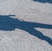 The image shows the lower body of a person wearing military camouflage uniform and tan combat boots walking on an asphalt path. A prominent shadow is cast on the pavement, revealing the silhouette of the full figure including what appears to be a helmet or headgear and possibly carried equipment. Green grass is visible along the edge of the path.