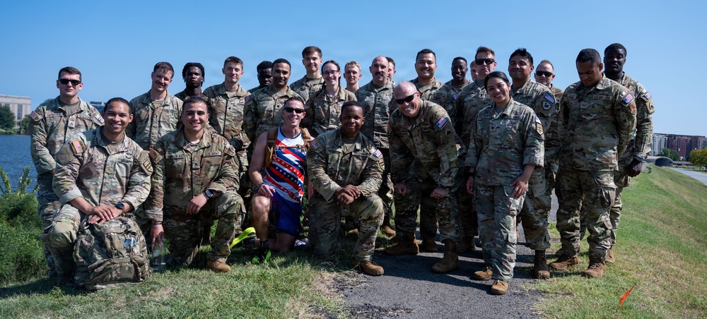 A large group of approximately 20-22 military personnel in U.S. Army camouflage (OCP) uniforms pose for a group photo outdoors near a body of water. Among them is one civilian wearing a patriotic-themed athletic racing outfit with a medal, suggesting a running or athletic event. The photo appears to be taken on a sunny day in an urban waterfront setting.