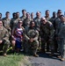 A large group of approximately 20-22 military personnel in U.S. Army camouflage (OCP) uniforms pose for a group photo outdoors near a body of water. Among them is one civilian wearing a patriotic-themed athletic racing outfit with a medal, suggesting a running or athletic event. The photo appears to be taken on a sunny day in an urban waterfront setting.