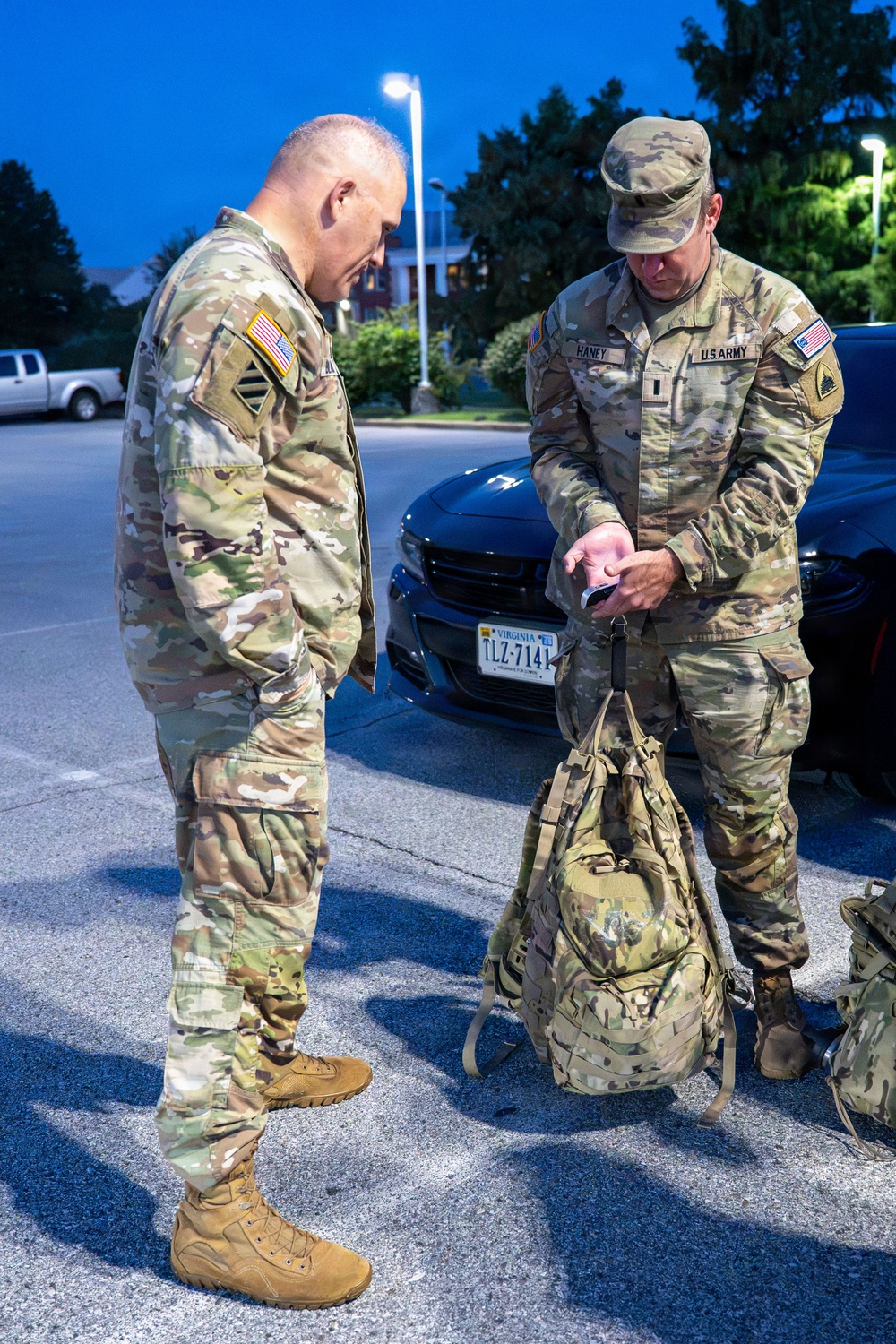 Two U.S. Army soldiers in Operational Camouflage Pattern (OCP) uniforms are standing in a parking lot during early morning or evening hours under artificial lighting. The soldier on the right, identified by name tape as 'HANEY,' is examining an item in his hand while standing over military-style rucksacks. Both soldiers appear to be preparing for or returning from a deployment or field exercise.