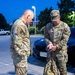 Two U.S. Army soldiers in Operational Camouflage Pattern (OCP) uniforms are standing in a parking lot during early morning or evening hours under artificial lighting. The soldier on the right, identified by name tape as 'HANEY,' is examining an item in his hand while standing over military-style rucksacks. Both soldiers appear to be preparing for or returning from a deployment or field exercise.