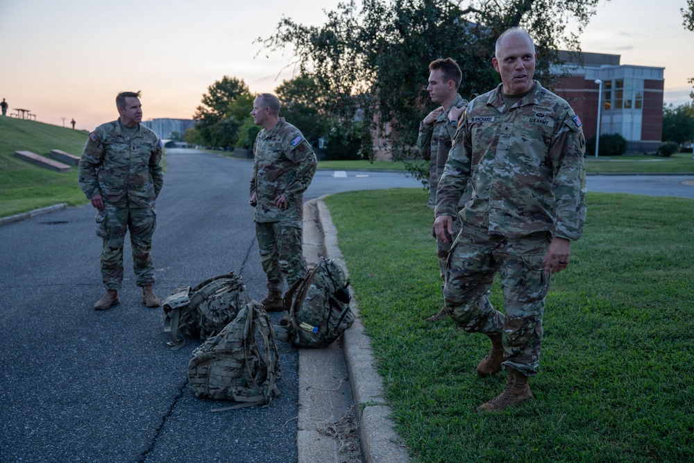 Four U.S. Army soldiers in OCP (Operational Camouflage Pattern) uniforms are gathered outdoors on a military installation at dawn or dusk. The soldier in the foreground has a visible name tape reading 'BLANCHARD' and a U.S. Army chest tape. Two military rucksacks are placed on the ground near the curb between the road and grass.