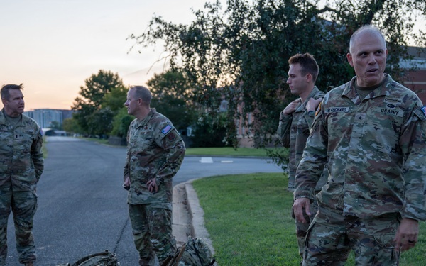 Four U.S. Army soldiers in OCP (Operational Camouflage Pattern) uniforms are gathered outdoors on a military installation at dawn or dusk. The soldier in the foreground has a visible name tape reading 'BLANCHARD' and a U.S. Army chest tape. Two military rucksacks are placed on the ground near the curb between the road and grass.