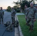 Four U.S. Army soldiers in OCP (Operational Camouflage Pattern) uniforms are gathered outdoors on a military installation at dawn or dusk. The soldier in the foreground has a visible name tape reading 'BLANCHARD' and a U.S. Army chest tape. Two military rucksacks are placed on the ground near the curb between the road and grass.