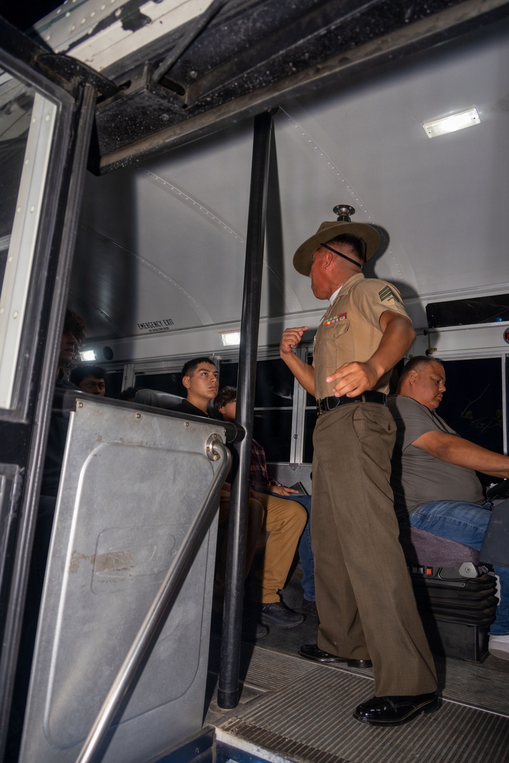 A uniformed military drill instructor, wearing a campaign cover (Smokey Bear hat) and tan uniform with ribbons, stands at the entrance of a bus addressing seated recruits or new enlistees. The scene appears to be a military reception or processing event, likely at night based on the dark exterior visible through the windows. The drill instructor is gesturing and appears to be giving instructions to the individuals seated on the bus.