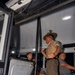 A uniformed military drill instructor, wearing a campaign cover (Smokey Bear hat) and tan uniform with ribbons, stands at the entrance of a bus addressing seated recruits or new enlistees. The scene appears to be a military reception or processing event, likely at night based on the dark exterior visible through the windows. The drill instructor is gesturing and appears to be giving instructions to the individuals seated on the bus.