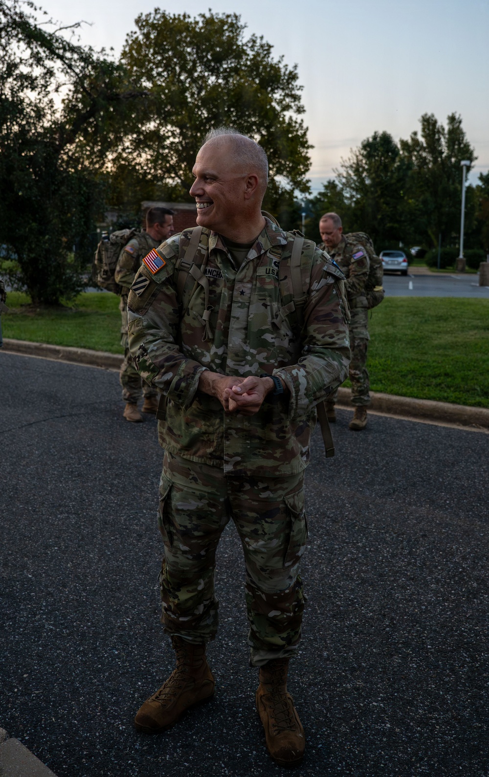 A smiling U.S. Army soldier in OCP (Operational Camouflage Pattern) uniform stands in the foreground on a paved surface, wearing a rucksack and displaying a one-star rank insignia indicating the rank of Brigadier General. Several other soldiers in similar uniforms with rucksacks are visible in the background. The setting appears to be a military installation during early morning or evening hours.