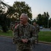 A smiling U.S. Army soldier in OCP (Operational Camouflage Pattern) uniform stands in the foreground on a paved surface, wearing a rucksack and displaying a one-star rank insignia indicating the rank of Brigadier General. Several other soldiers in similar uniforms with rucksacks are visible in the background. The setting appears to be a military installation during early morning or evening hours.