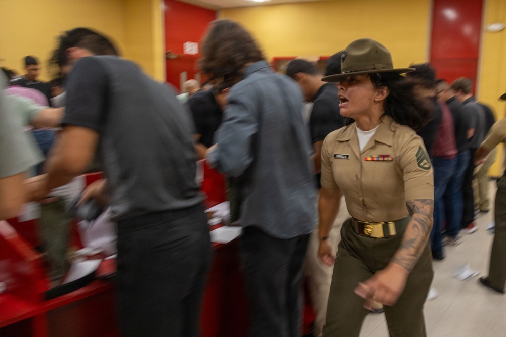 A female U.S. Marine drill instructor in tan service uniform and campaign cover (drill sergeant hat) is actively shouting commands at a group of recruits or civilians in what appears to be a processing or reception facility. The scene captures a high-energy, chaotic moment with motion blur indicating rapid movement among the group. The setting features yellow walls and red fixtures consistent with a military processing station or MEPS (Military Entrance Processing Station).
