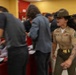 A female U.S. Marine drill instructor in tan service uniform and campaign cover (drill sergeant hat) is actively shouting commands at a group of recruits or civilians in what appears to be a processing or reception facility. The scene captures a high-energy, chaotic moment with motion blur indicating rapid movement among the group. The setting features yellow walls and red fixtures consistent with a military processing station or MEPS (Military Entrance Processing Station).