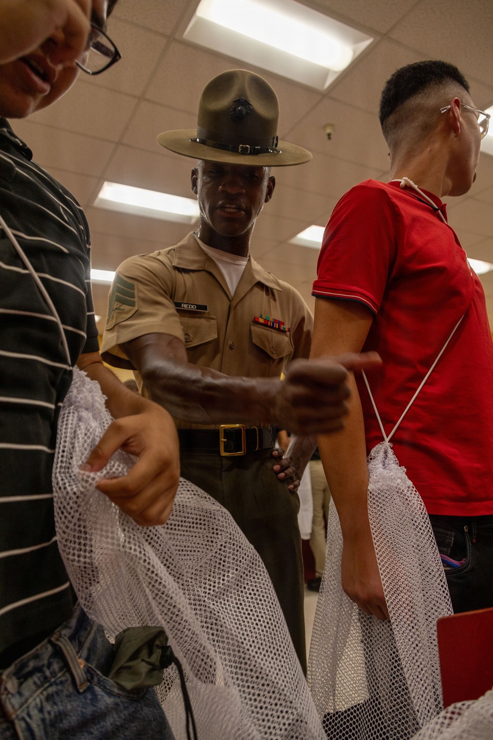 A U.S. Marine Corps drill instructor wearing a campaign cover (drill sergeant hat) and tan uniform with the name tape 'REDD' is interacting with what appear to be new recruits in an indoor facility. The recruits are holding white mesh laundry bags, suggesting this is a processing or receiving stage of basic training. The setting appears to be a recruit receiving or supply area on a Marine Corps base.