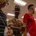 A U.S. Marine Corps drill instructor wearing a campaign cover (drill sergeant hat) and tan uniform with the name tape 'REDD' is interacting with what appear to be new recruits in an indoor facility. The recruits are holding white mesh laundry bags, suggesting this is a processing or receiving stage of basic training. The setting appears to be a recruit receiving or supply area on a Marine Corps base.