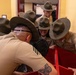 A group of U.S. Marine Corps drill instructors in tan service uniforms and campaign covers (drill instructor hats) are clustered together in what appears to be an indoor military facility. One drill instructor in the center appears to be shouting with an intense expression, while others lean in around a red object in the foreground. The scene suggests a training or instructional exercise environment.