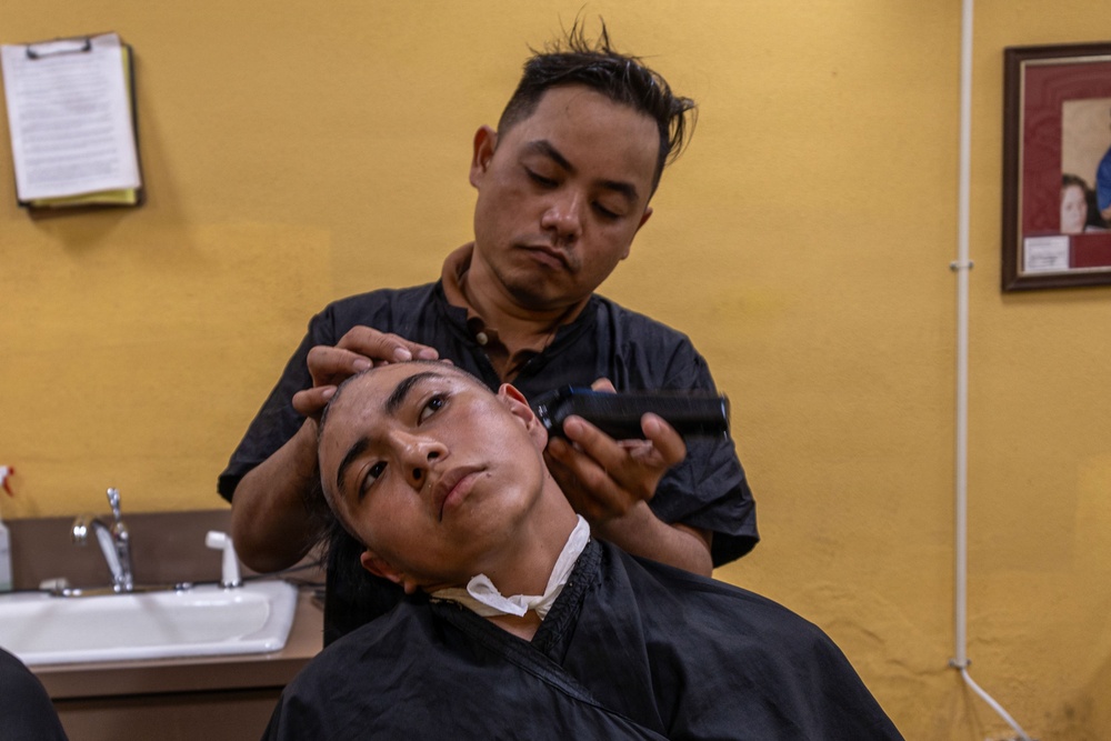 A barber is cutting the hair of a young male client in a barbershop setting. The barber, dressed in a black shirt, holds electric clippers and positions the client's head with his other hand. The client is draped in a black barber cape and appears relaxed.