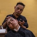 A barber is cutting the hair of a young male client in a barbershop setting. The barber, dressed in a black shirt, holds electric clippers and positions the client's head with his other hand. The client is draped in a black barber cape and appears relaxed.