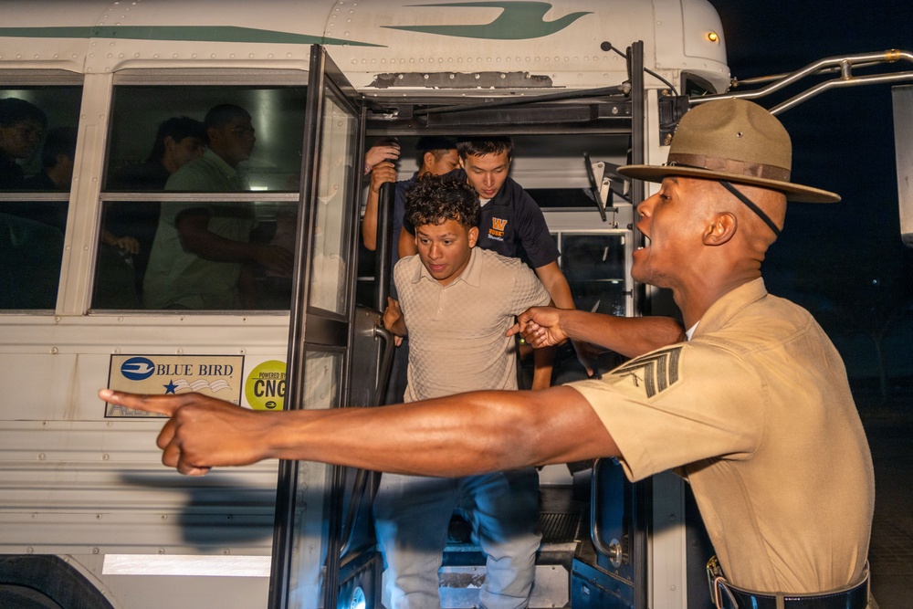 A drill instructor or military/law enforcement officer in a campaign cover (Smokey Bear hat) and tan uniform is directing recruits or trainees off a Blue Bird CNG-powered bus at night. The officer is pointing emphatically while recruits exit the bus in an urgent manner. This appears to be a military or law enforcement training scenario, consistent with a boot camp or recruit processing arrival.