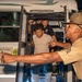 A drill instructor or military/law enforcement officer in a campaign cover (Smokey Bear hat) and tan uniform is directing recruits or trainees off a Blue Bird CNG-powered bus at night. The officer is pointing emphatically while recruits exit the bus in an urgent manner. This appears to be a military or law enforcement training scenario, consistent with a boot camp or recruit processing arrival.