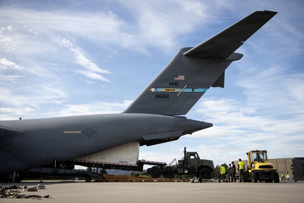 A U.S. Air Force C-17 Globemaster III cargo aircraft assigned to Dover Air Force Base (tail number 66166, AMC) is shown on a flight line during a cargo loading or unloading operation. Ground personnel and military vehicles, including a forklift and a military truck, are positioned near the rear of the aircraft. The scene appears to be at a military airfield under partly cloudy skies.