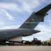 A U.S. Air Force C-17 Globemaster III cargo aircraft assigned to Dover Air Force Base (tail number 66166, AMC) is shown on a flight line during a cargo loading or unloading operation. Ground personnel and military vehicles, including a forklift and a military truck, are positioned near the rear of the aircraft. The scene appears to be at a military airfield under partly cloudy skies.
