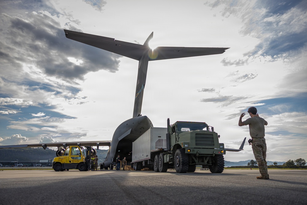 A military cargo aircraft, likely a C-17 Globemaster III, is being loaded or unloaded on an airfield tarmac. A military tactical truck with a trailer is positioned at the rear cargo ramp of the aircraft, while ground crew personnel and a yellow forklift assist with the operation. A soldier in camouflage uniform stands to the right, appearing to direct or signal the vehicle.