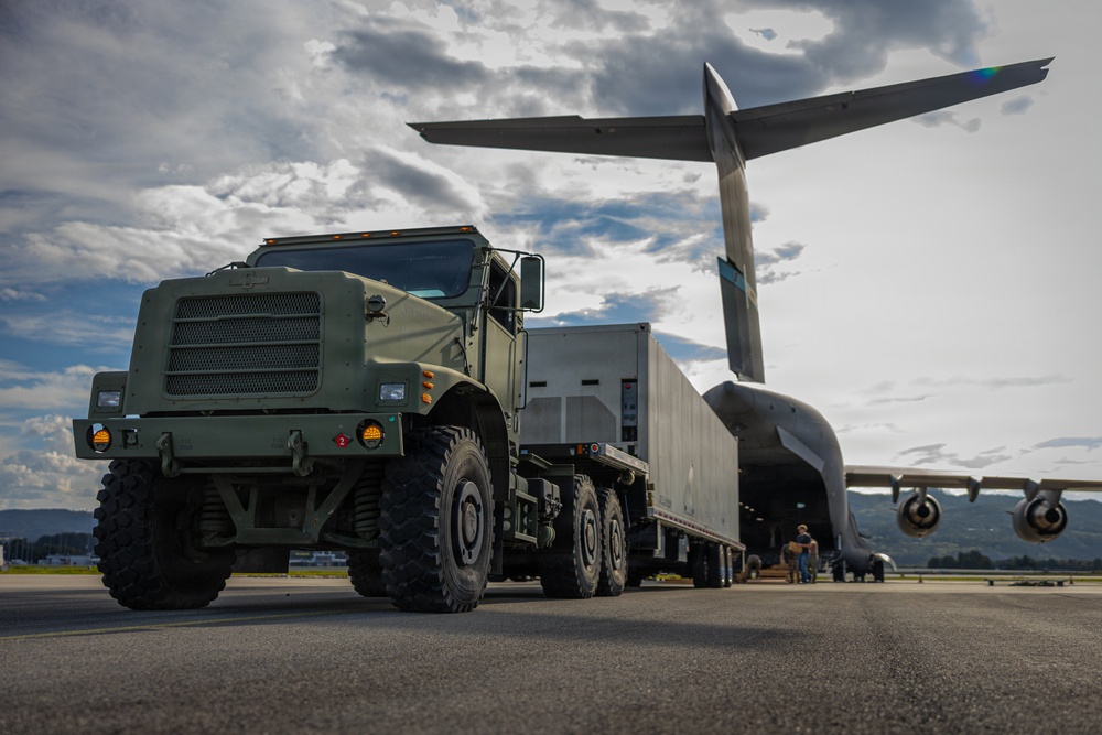 A military heavy-duty truck in olive drab green is pulling a flatbed trailer loaded with cargo out of or into a large military transport aircraft. The scene takes place on an airfield tarmac under a partly cloudy sky. The low-angle perspective emphasizes the scale of both the vehicle and the aircraft.