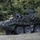 A military armored wheeled vehicle, consistent with a Stryker-type 8x8 armored personnel carrier, is positioned at the edge of a wooded treeline. The vehicle is draped with a woodland camouflage net and has a mounted weapon system visible on top. One soldier in combat uniform and helmet is seated atop the vehicle, appearing to observe the surrounding area.