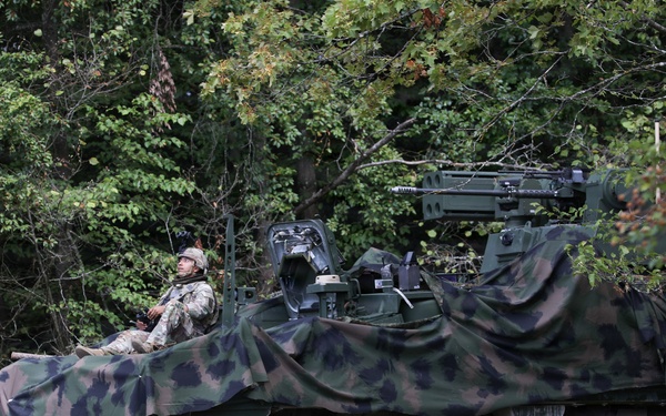 A military armored wheeled vehicle, consistent with a Stryker-type 8x8 armored personnel carrier, is positioned at the edge of a wooded treeline. The vehicle is draped with a woodland camouflage net and has a mounted weapon system visible on top. One soldier in combat uniform and helmet is seated atop the vehicle, appearing to observe the surrounding area.