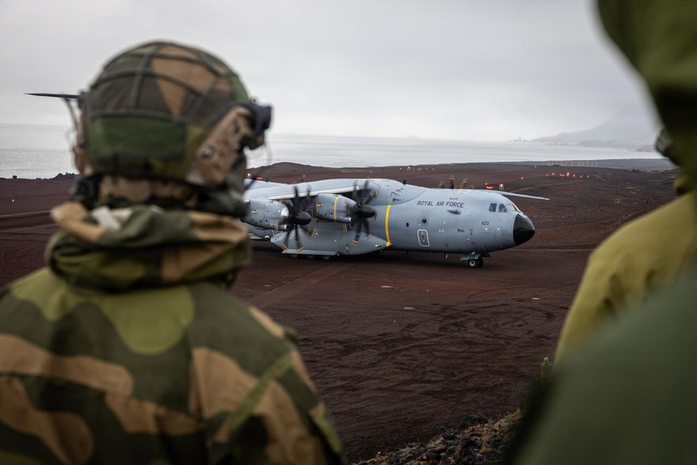 Two soldiers in camouflage uniforms and helmets observe a Royal Air Force Airbus A400M Atlas military transport aircraft (tail number 420) on a remote airstrip. The terrain appears volcanic or arctic, with dark reddish-brown ground and a body of water visible in the background under an overcast sky. This appears to be a military exercise or operational deployment in a remote northern or arctic location.