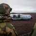 Two soldiers in camouflage uniforms and helmets observe a Royal Air Force Airbus A400M Atlas military transport aircraft (tail number 420) on a remote airstrip. The terrain appears volcanic or arctic, with dark reddish-brown ground and a body of water visible in the background under an overcast sky. This appears to be a military exercise or operational deployment in a remote northern or arctic location.