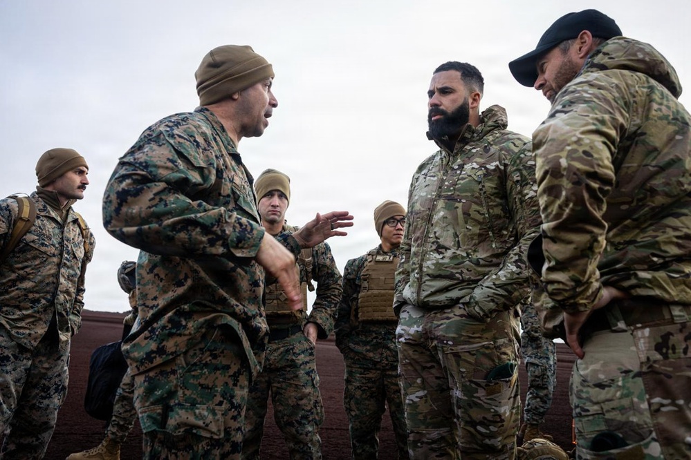A group of military personnel in two distinct camouflage patterns are engaged in a briefing or discussion outdoors in overcast conditions. One individual in MARPAT (Marine pattern) camouflage appears to be speaking and gesturing to others wearing MultiCam/OCP pattern uniforms, suggesting a joint or inter-service interaction. The setting appears to be a training environment or operational staging area.