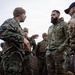 A group of military personnel in two distinct camouflage patterns are engaged in a briefing or discussion outdoors in overcast conditions. One individual in MARPAT (Marine pattern) camouflage appears to be speaking and gesturing to others wearing MultiCam/OCP pattern uniforms, suggesting a joint or inter-service interaction. The setting appears to be a training environment or operational staging area.