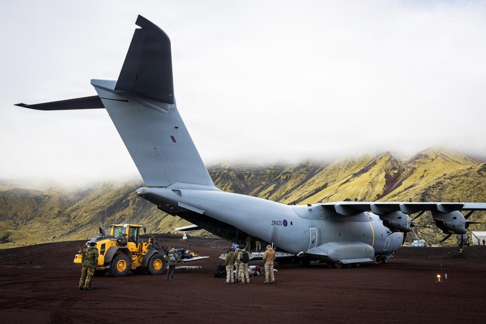 A Royal Air Force Airbus A400M Atlas military transport aircraft with tail code 'ZM420' is parked on a dark volcanic terrain, likely in Iceland or a similar volcanic landscape. Ground crew and military personnel are conducting loading or unloading operations using a yellow front-end loader/wheel loader. The aircraft's rear cargo ramp appears to be open as personnel work around the aft section.