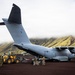 A Royal Air Force Airbus A400M Atlas military transport aircraft with tail code 'ZM420' is parked on a dark volcanic terrain, likely in Iceland or a similar volcanic landscape. Ground crew and military personnel are conducting loading or unloading operations using a yellow front-end loader/wheel loader. The aircraft's rear cargo ramp appears to be open as personnel work around the aft section.