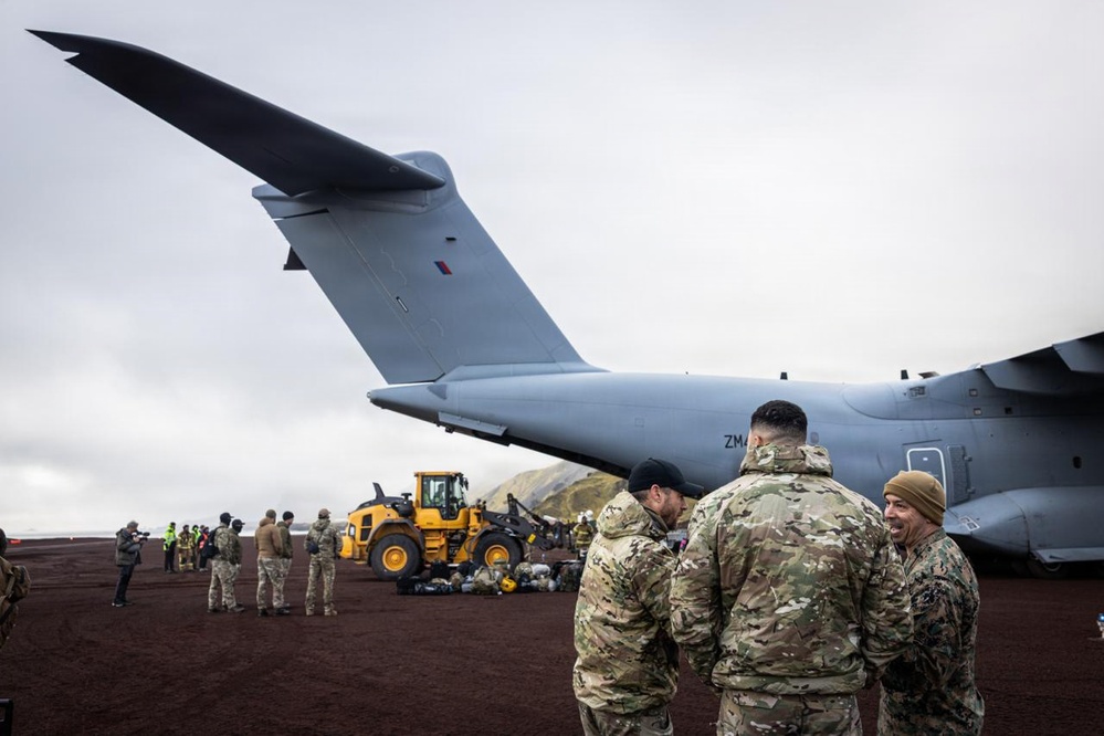 Military personnel are gathered on a dark volcanic or gravel airstrip near a large grey military transport aircraft, identifiable by the partial tail registration 'ZM' and a Union Jack roundel, consistent with a Royal Air Force Airbus A400M Atlas. Ground support equipment including a yellow front-end loader is visible, and cargo or equipment appears to be offloaded or staged nearby. The overcast sky and dark terrain suggest a remote or austere operating environment.