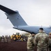 Military personnel are gathered on a dark volcanic or gravel airstrip near a large grey military transport aircraft, identifiable by the partial tail registration 'ZM' and a Union Jack roundel, consistent with a Royal Air Force Airbus A400M Atlas. Ground support equipment including a yellow front-end loader is visible, and cargo or equipment appears to be offloaded or staged nearby. The overcast sky and dark terrain suggest a remote or austere operating environment.