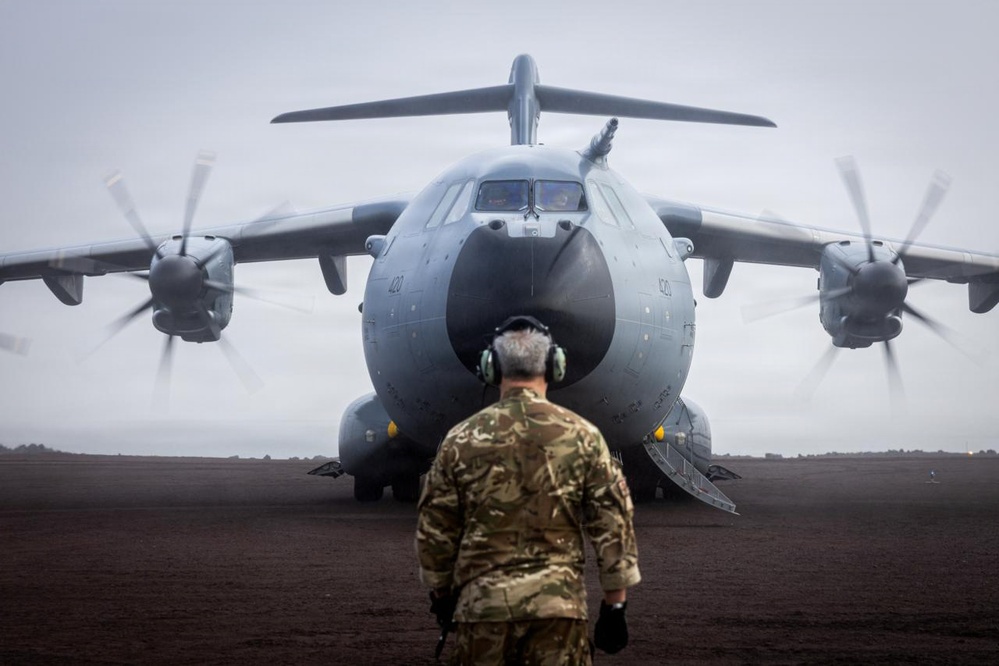 A military ground crew member wearing camouflage uniform and ear protection stands facing a large military transport aircraft with spinning propellers on a dark tarmac or unpaved surface. The aircraft appears to be an Airbus A400M Atlas turboprop military transport, identifiable by its distinctive nose shape, four turboprop engines with eight-bladed propellers, and grey livery. The overcast, misty conditions and front-on perspective create a dramatic composition.
