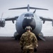 A military ground crew member wearing camouflage uniform and ear protection stands facing a large military transport aircraft with spinning propellers on a dark tarmac or unpaved surface. The aircraft appears to be an Airbus A400M Atlas turboprop military transport, identifiable by its distinctive nose shape, four turboprop engines with eight-bladed propellers, and grey livery. The overcast, misty conditions and front-on perspective create a dramatic composition.