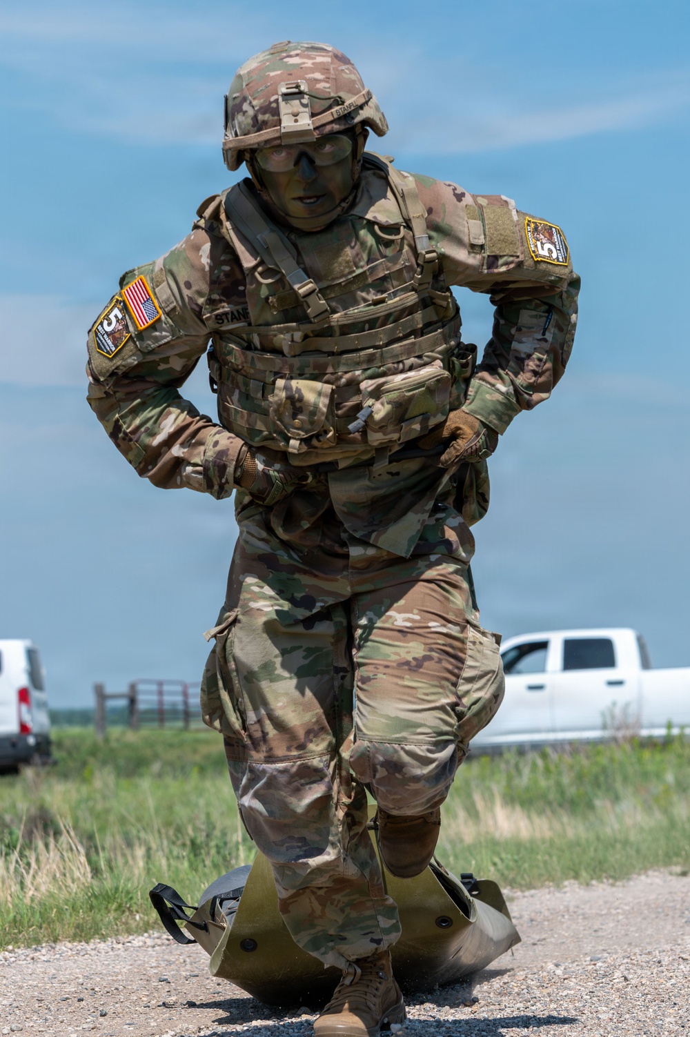 A U.S. Army soldier in full OCP (Operational Camouflage Pattern) uniform and body armor is dragging a litter or equipment bag along a gravel road. The soldier appears to be participating in a physical fitness or combat skills competition, wearing tactical gear including a helmet, plate carrier, and protective eyewear. Patches on both shoulders indicate unit affiliation, including a U.S. flag and what appears to be a '5th' unit insignia.