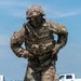 A U.S. Army soldier in full OCP (Operational Camouflage Pattern) uniform and body armor is dragging a litter or equipment bag along a gravel road. The soldier appears to be participating in a physical fitness or combat skills competition, wearing tactical gear including a helmet, plate carrier, and protective eyewear. Patches on both shoulders indicate unit affiliation, including a U.S. flag and what appears to be a '5th' unit insignia.