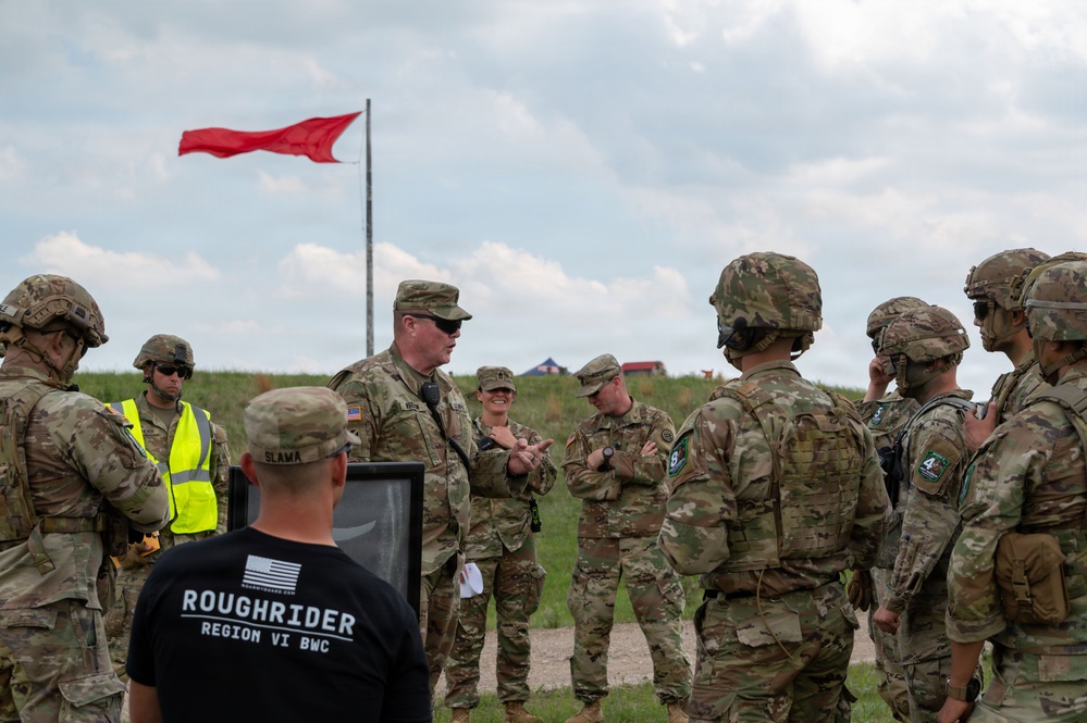A group of military personnel in OCP (Operational Camouflage Pattern) uniforms are gathered in an outdoor field setting, appearing to receive a briefing or debriefing. A red range flag is visible flying on a pole in the background, indicating an active or recently active firing range. One civilian in a black 'Roughrider Region VI BWC' t-shirt is also present among the group.